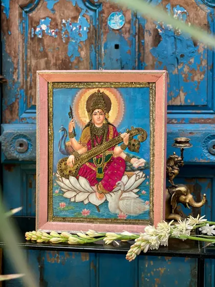 The Goddess Saraswati cloth oleograph, beautifully styled against a vintage blue door with flowers. This shows how these art pieces can become the centerpiece of an elegant, culturally-rooted display.