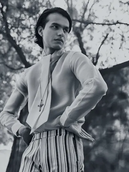A black and white editorial shot for Kaltblut Magazine. The androgynous styling, featuring a sheer shirt with a built-in scarf, striped trousers, and a cross pendant, explores themes of gender fluidity in high fashion.