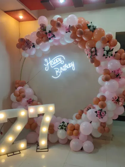 A chic 27th birthday decoration with a balloon ring in pastel pink and rose gold, a neon sign, and large marquee numbers.