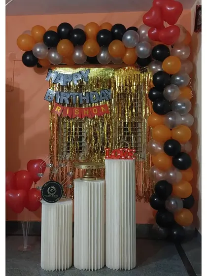 Even a simple corner can become a festive photo booth. This setup uses a gold shimmer curtain and a balloon garland in orange, black, and silver, along with pedestals for the cake, to maximize a small space.