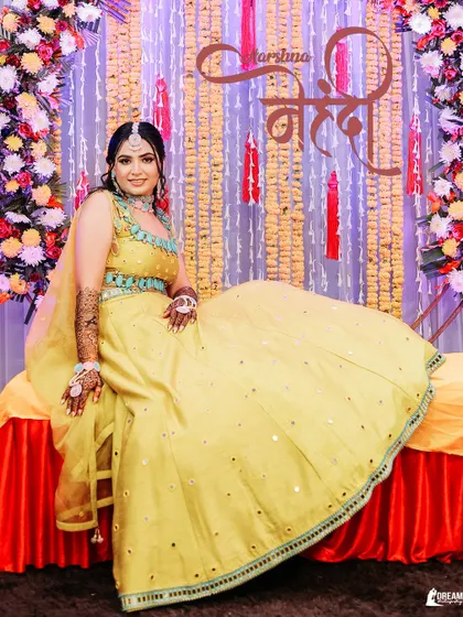 A full-length portrait of bride Harshna at her Mehndi, seated against a vibrant backdrop of flowers, showcasing her bright yellow lehenga.