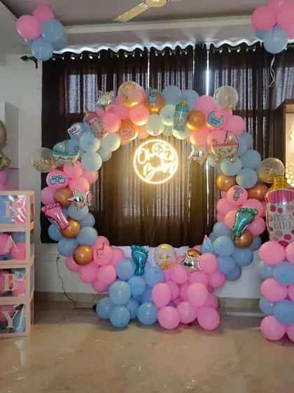 A gender reveal or baby shower decoration with a mix of pink and blue balloons. The setup includes a balloon ring, a neon sign, and "BABY" blocks.