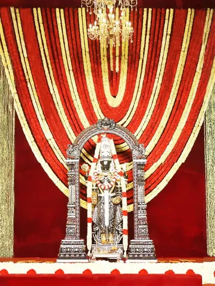 A stunning backdrop of red and gold floral garlands frames the sacred deity. Paired with a glowing chandelier, this design radiates divine energy and creates an atmosphere of pure elegance and grace.