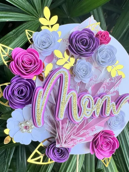 Our stunning handmade floral "Mom" topper for Mother's Day. This intricate piece features paper flowers, gold leaves, and a glittery, layered "Mom" script.