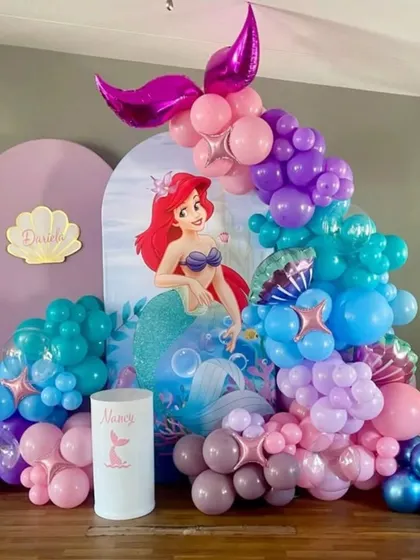 A magical Little Mermaid theme for a birthday party, featuring an Ariel backdrop and a balloon arrangement in shades of purple, pink, and teal to mimic the ocean.