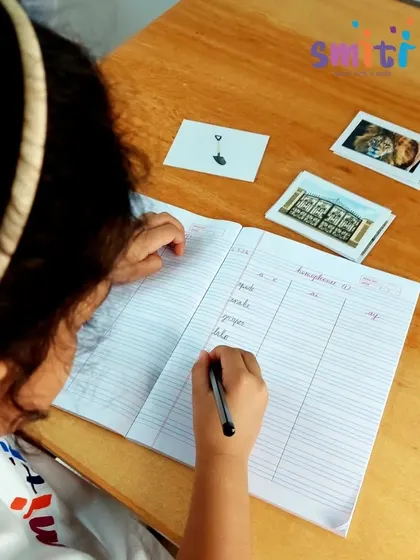 A student writes words in her notebook, matching them to picture cards. This activity connects phonetic sounds with written symbols, forming the basis of reading and writing.