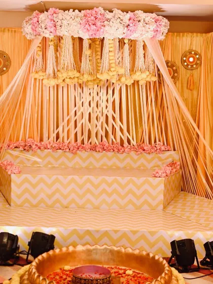 Here is the full view of the beautiful Haldi ceremony stage I designed. The setup features a canopy of hanging floral strings, a tiered seating area with a chevron pattern, and a large brass uruli in the front for the rituals.