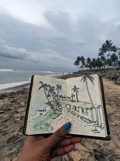My sketchbook held up against the cloudy sky and coconut trees of Varkala beach. The pen and watercolor sketch captures the iconic cliffside path and the sea.
