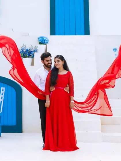 A dramatic and colorful pre-wedding shot. The long red trail of her infinity gown creates a stunning visual against the bright blue and white background.