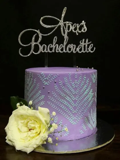 This purple bachelorette cake showcases a beautiful stenciling technique. The delicate silver leaf pattern on the side is complemented by a fresh white rose and a glittery custom cake topper.