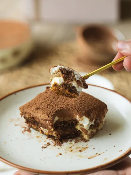 A perfect slice of our Classic Tiramisu on a plate. You can see the delicate layers of espresso-soaked biscuits and velvety mascarpone cream, finished with a dusting of cocoa powder.