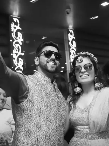 A joyful black and white photo of the couple dancing at their Haldi party. Their hands are in the air, and they are surrounded by smiling family members.