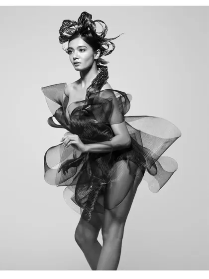 A black and white portrait from the "HAIR" series, where the model is enveloped in a dress made of sculpted mesh. The image plays with transparency, structure, and form.