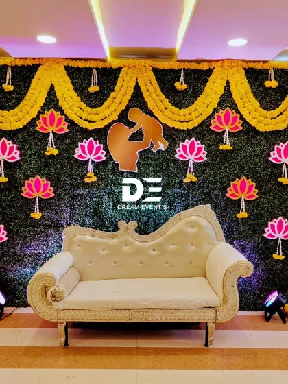 A simple yet charming baby shower backdrop with a green foliage wall, marigold garlands, and lotus cutouts. The elegant chaise lounge provides a perfect spot for the mother-to-be.