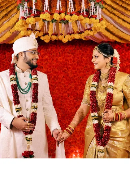 A candid photo of the couple holding hands and smiling at each other, standing before a vibrant red and yellow floral backdrop.
