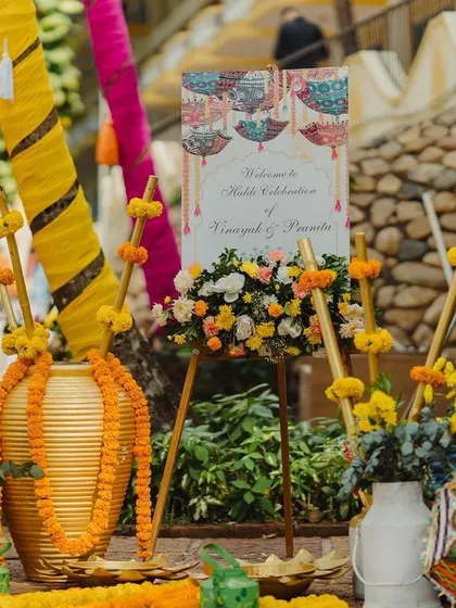 A colorful and quirky entrance to a Haldi Carnival, with a welcome sign, traditional umbrellas, and vibrant floral arrangements.