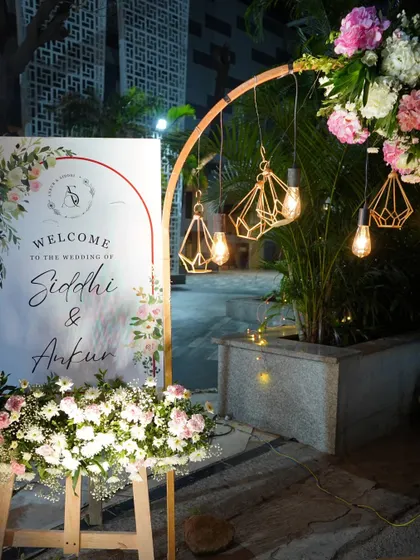 This welcome sign for Siddhi and Ankur is framed by an elegant wooden arch and lit by geometric hanging lamps. The design is modern, warm, and personal, perfect for an evening celebration.