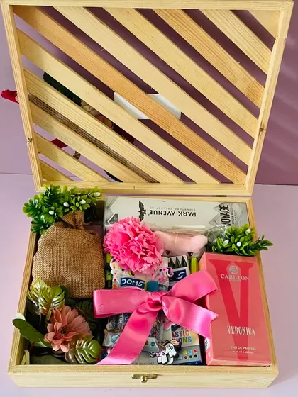 The open pinewood crate reveals a variety of gifts, including perfumes, snacks, and activities, all tied together with a beautiful pink ribbon.