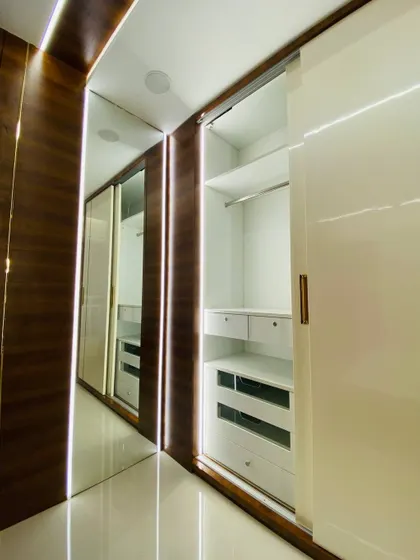 This is a walk-in wardrobe with glossy white sliding doors and a rich wood frame. The integrated profile lights along the side not only look great but also help in illuminating the wardrobe's contents.