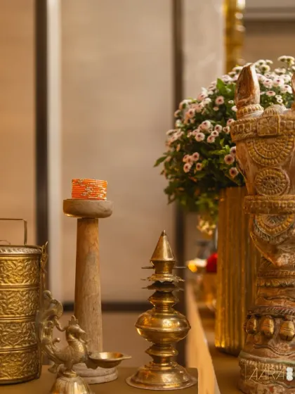 A still life of traditional decor elements, including a vintage brass tiffin carrier, a hand-carved wooden pillar, and an antique lamp. These pieces are carefully styled to create vignettes that tell a story of heritage and craftsmanship.