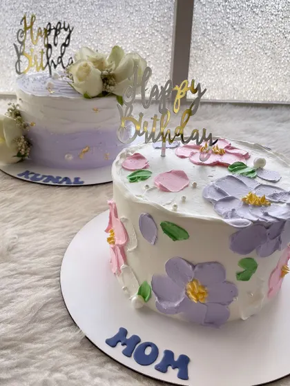 A pair of pretty pastel cakes. One is a simple floral design, while the other is decorated with fresh white roses and a hint of lilac.