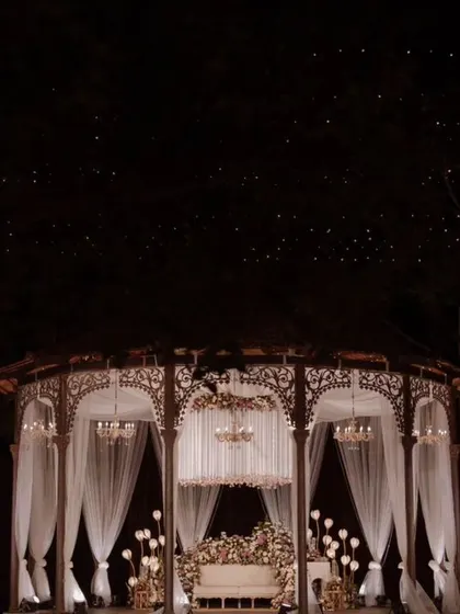A grand view of a reception stage set within a rustic gazebo, transformed into an elegant space with white drapes, chandeliers, and overflowing florals.