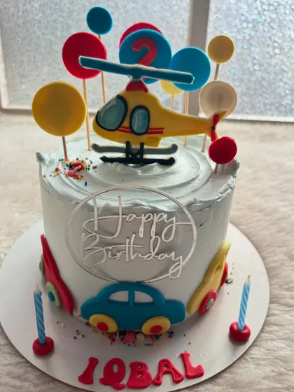 A transportation-themed cake featuring a helicopter and cars. The bright primary colors and fun toppers make it a great choice for a second birthday.