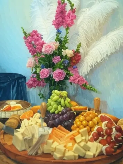 An artistic, painted-effect image of a lavish cheese board. The platter is adorned with a stunning floral arrangement and features a colorful assortment of cheeses, grapes, and caprese skewers.