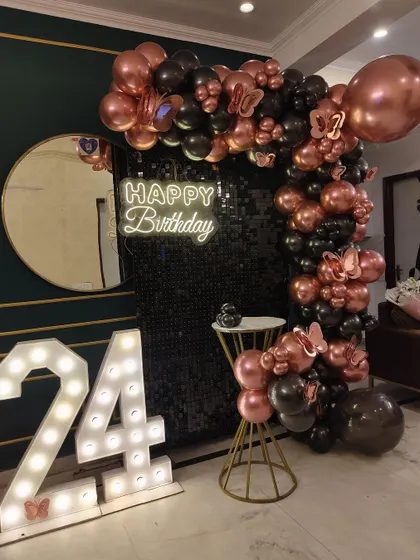 A chic 24th birthday setup with a black shimmer wall. The decor features a rose gold and black balloon arch and large marquee numbers.