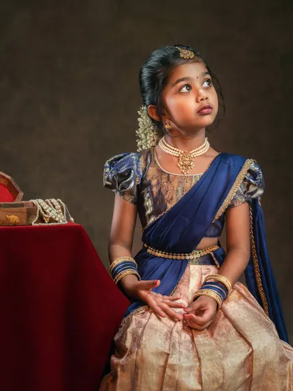 A girl in a traditional blue and gold lehenga looks up thoughtfully. The classic lighting and pose give this portrait a timeless, painterly quality.
