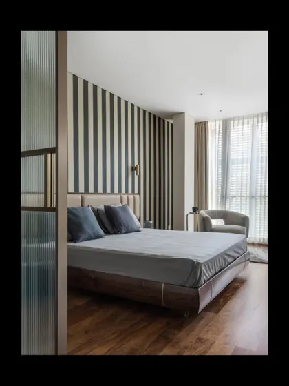 This bedroom is a study in refined comfort, featuring a bold striped wallpaper that adds graphic interest without overwhelming the space. A fluted glass partition provides a subtle separation, enhancing privacy while allowing light to filter through, creating a serene and layered retreat.