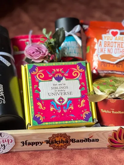 This Rakhi hamper for a brother is curated with a personalized sipper bottle, snacks, and a fun "Siblings in every universe" box, all arranged in a neat wooden tray.