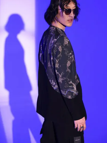 This shot from a menswear lookbook features a jacket with a graphic print panel against a vibrant blue background. The styling is edgy and modern, highlighting the unique design details of the garment.