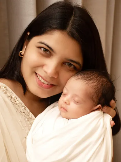 A glowing new mother smiles warmly at the camera while holding her peacefully sleeping newborn, a simple and beautiful portrait of the maternal bond.