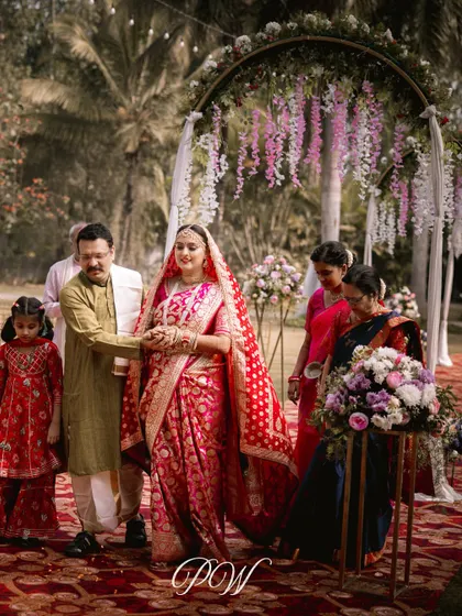 The bride's grand entrance, walking down an aisle flanked by family and beautiful floral arrangements. The path leads to a dreamy mandap with hanging wisteria, setting the stage for a magical ceremony.