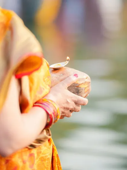A woman holds a coconut with a lit diya on top, a key part of the Chhath Puja offering. The light symbolizes the dispelling of darkness.