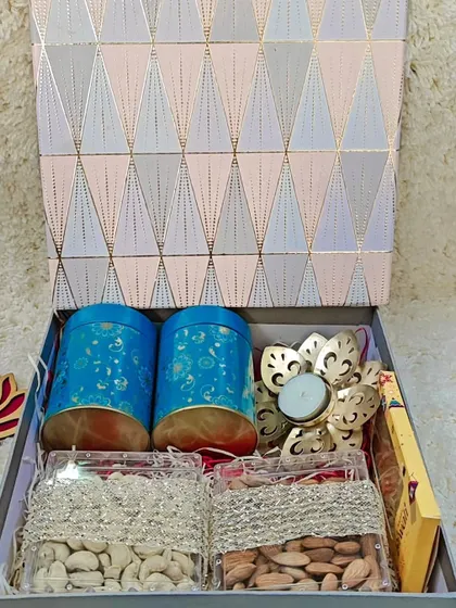 This hamper combines health and indulgence. It features two decorative tins, boxes of premium almonds and cashews, and a lotus-shaped brass diya, all in a stylish geometric-print box.