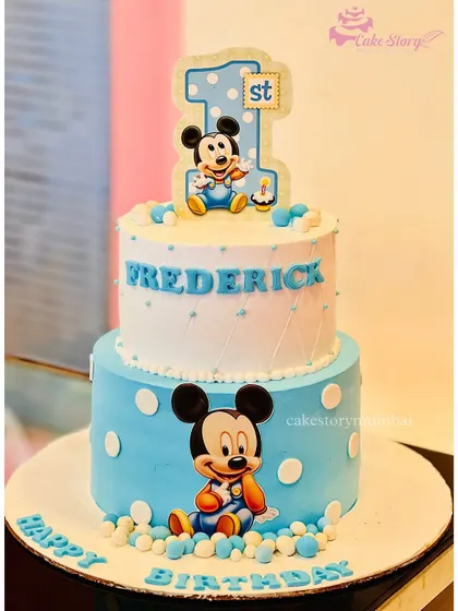 A classic Mickey Mouse first birthday cake. This two-tier design in blue and white is decorated with polka dots and adorable illustrations of baby Mickey.