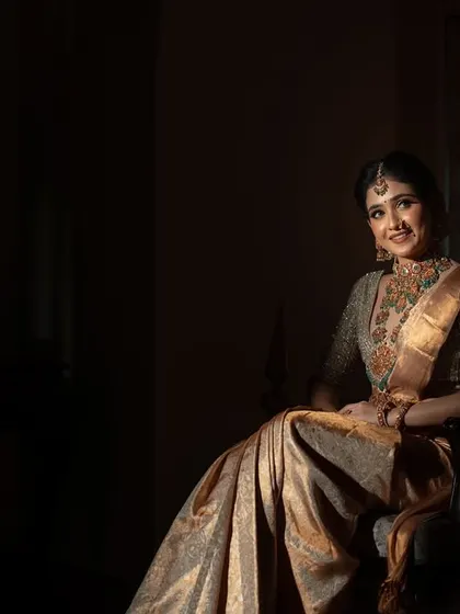 A seated portrait of the bride, with a single light source creating a dramatic and elegant mood. The shadows emphasize her form and the richness of her golden saree.
