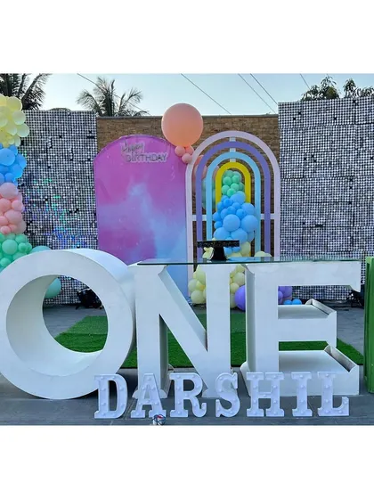 A contemporary outdoor first birthday setup with a large "ONE" table, colorful props, and shimmer wall panels against a stone wall.