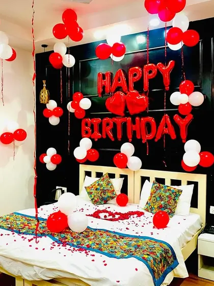 A perfect, straight-on shot of the romantic red and white birthday bedroom decoration, showcasing the entire setup from the foil balloons to the petals on the bed.