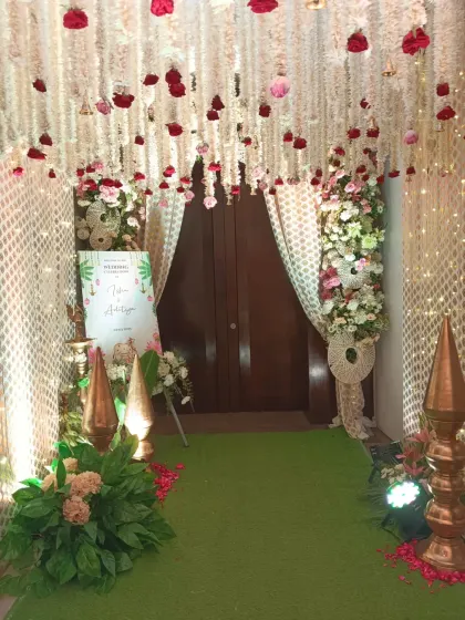 An entrance that blends tradition with elegance. Guests are welcomed through a pathway lined with a green carpet and traditional decor elements, under a ceiling of hanging white and red floral strings.