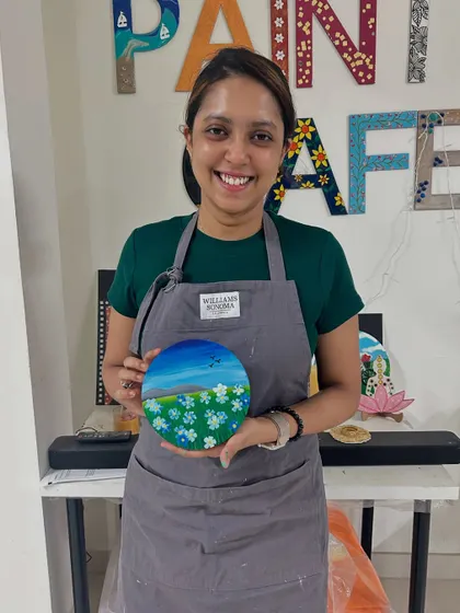 A smiling participant in an apron holds her circular canvas painting of a field of flowers.
