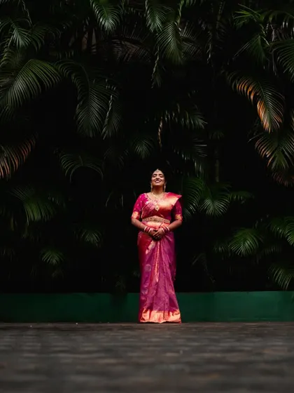 A striking portrait of a bride in a pink saree against a dark wall of foliage. The composition is simple yet powerful.