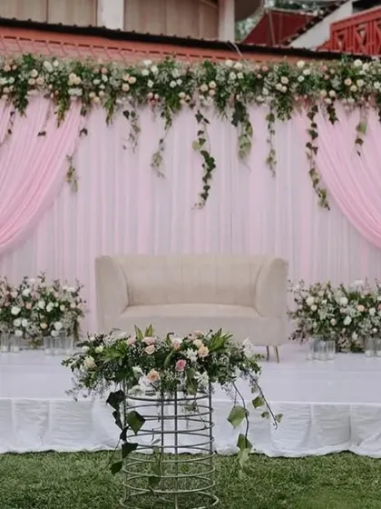 A soft and romantic stage setup perfect for an outdoor or daytime reception. The design uses flowing pink drapery, a clean white platform, and abundant greenery with white and pink florals for a fresh, whimsical feel.