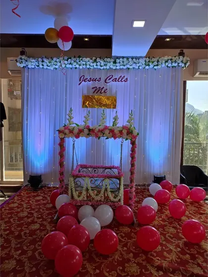 A closer look at the cradle decorated with artificial flowers for the naming ceremony. The setup is placed on a red carpeted stage, making it the focal point of the event.