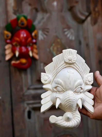 A white Ganesha mask, hand-carved and ready for painting, shown next to a finished red one. This highlights the process and the craftsmanship that goes into each unique piece of Nepalese folk art.