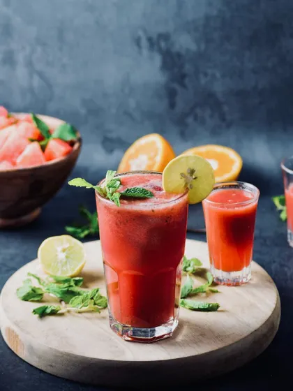 A refreshing watermelon and mint juice, styled with fresh fruit on a wooden board. The dark, moody background makes the bright red color of the drink pop.