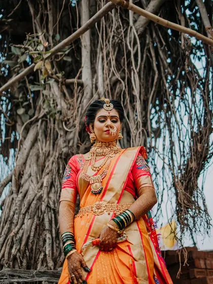A powerful portrait of a Maharashtrian bride in a stunning orange saree, standing before a large banyan tree.