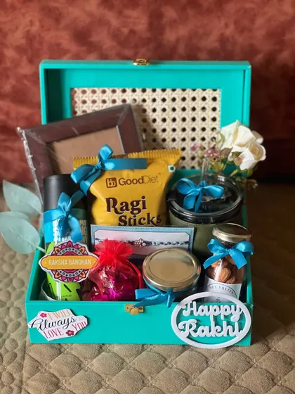 A stylish Rakhi hamper in a teal trunk box with cane detailing. It includes a photo frame, healthy snacks, and a "Happy Rakhi" tag.